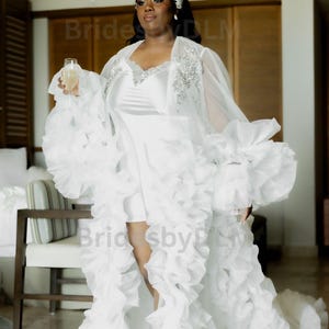 May include: A white bridal robe with a short dress underneath. The robe has a ruffled design with a sheer overlay on the shoulders and sleeves. The dress is a simple white satin. The bride is holding a champagne glass.
