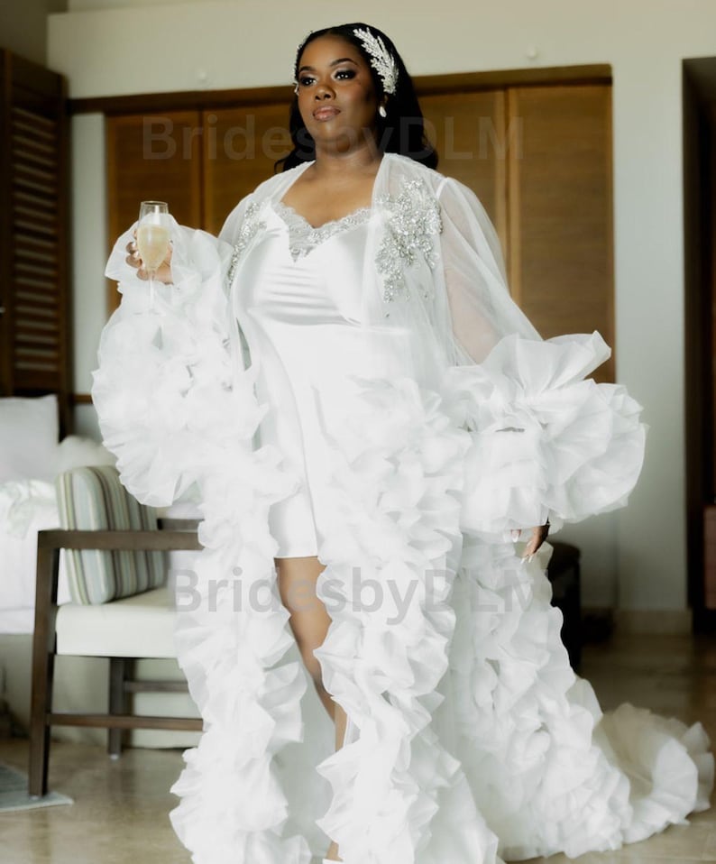 May include: A white bridal robe with a short dress underneath. The robe has a ruffled design with a sheer overlay on the shoulders and sleeves. The dress is a simple white satin. The bride is holding a champagne glass.