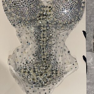 May include: A white bodysuit covered in silver rhinestones and pearls. The bodice is heavily embellished with the sparkling stones, while the lower portion features a sheer, delicate fabric. The garment is displayed on a white surface.