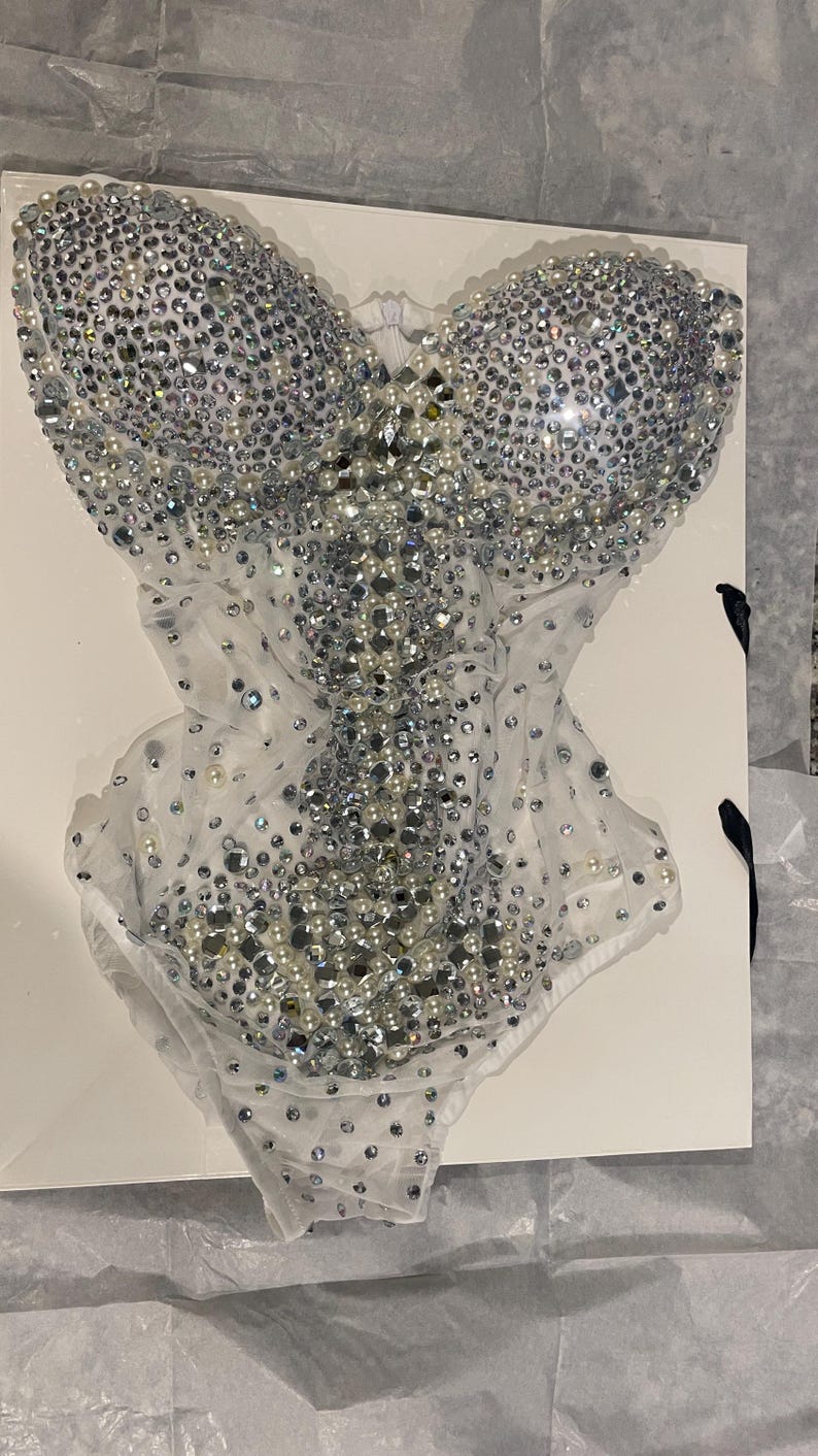 May include: A white bodysuit covered in silver rhinestones and pearls. The bodice is heavily embellished with the sparkling stones, while the lower portion features a sheer, delicate fabric. The garment is displayed on a white surface.