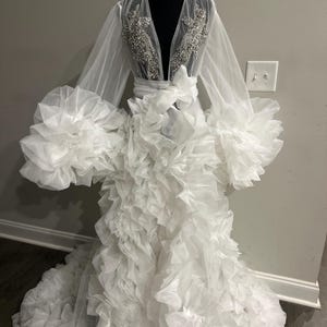 May include: A white bridal robe displayed on a mannequin. The robe features long, sheer sleeves with large ruffled cuffs. The bodice has a deep V-neckline with intricate silver embellishments. The skirt is voluminous, cascading with layers of ruffled fabric, creating a dramatic effect.