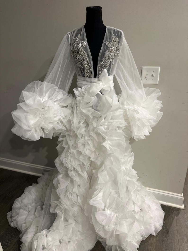 May include: A white bridal robe displayed on a mannequin. The robe features long, sheer sleeves with large ruffled cuffs. The bodice has a deep V-neckline with intricate silver embellishments. The skirt is voluminous, cascading with layers of ruffled fabric, creating a dramatic effect.
