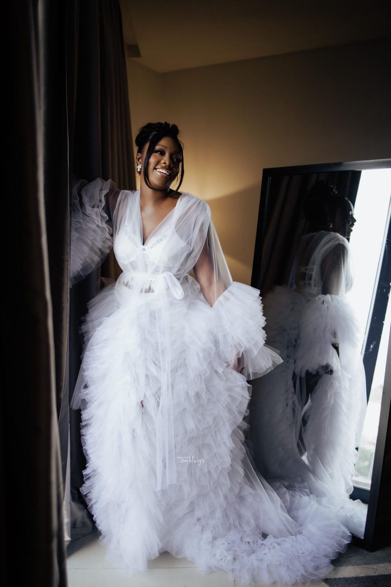 May include: A white tulle robe with ruffled layers and long sleeves. The robe is tied at the waist with a white ribbon. The robe is sheer and has a delicate lace trim.
