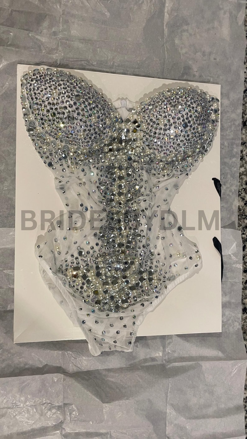 May include: A silver bodysuit covered in sparkling rhinestones and pearls. The garment is on a white surface, with the text "BRIDE" and "YDLM" visible. The bodysuit has a sheer, white mesh bottom.