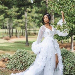 May include: A white tulle robe with a ruffled skirt and long sleeves. The robe has a satin bodice and a sash tied at the waist. The robe is worn by a person with dark hair and a tiara.