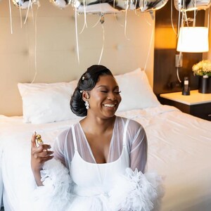 May include: A woman wearing a white tulle robe with ruffled sleeves. The robe is sheer and has a white slip underneath. The woman is smiling and holding a small bottle of perfume.