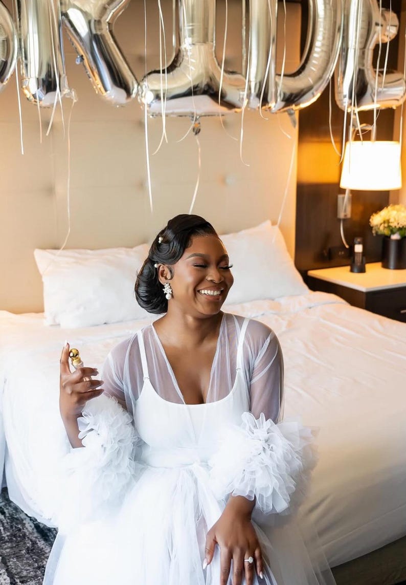 May include: A woman wearing a white tulle robe with ruffled sleeves. The robe is sheer and has a white slip underneath. The woman is smiling and holding a small bottle of perfume.