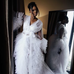 May include: A white tulle robe with ruffled sleeves and a long train. The robe is tied at the waist with a white sash. The robe is sheer and has a delicate, feminine look.