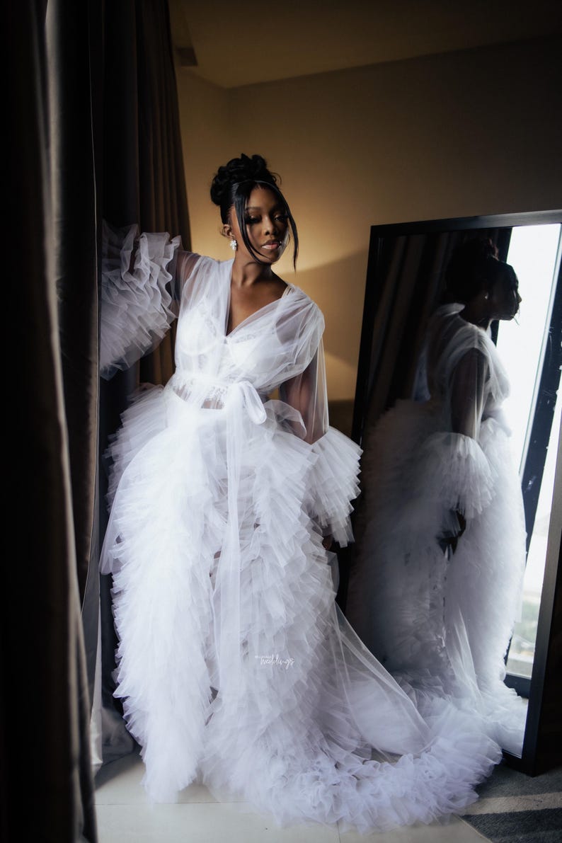 May include: A white tulle robe with ruffled sleeves and a long train. The robe is tied at the waist with a white sash. The robe is sheer and has a delicate, feminine look.