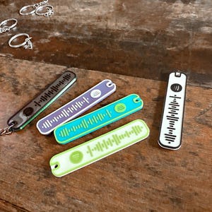 Könnte beinhalten: Vier verschiedene Schlüsselanhänger mit einem schwarz-weißen Soundwave-Design. Die Schlüsselanhänger sind aus Kunststoff und in verschiedenen Farben erhältlich, darunter Schwarz, Weiß, Grün, Blau und Lila.