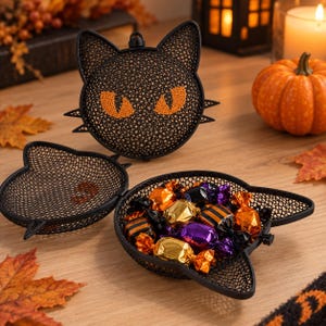 Könnte beinhalten: Ein schwarzer Metallgitterbehälter in Form eines Katzenkopfes mit orangefarbenen Augen. Der Behälter ist aufklappbar und enthält eine Auswahl an Süßigkeiten, die in glänzende orangefarbene, violette und schwarze Folie eingewickelt sind.