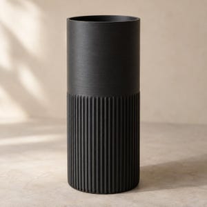 Könnte beinhalten: Eine hohe, zylindrische schwarze Vase mit einem glatten oberen Bereich und einem vertikal gerippten, strukturierten unteren Teil. Das Gefäß hat eine weite Öffnung und einen flachen Boden, gefertigt mit einem matten Finish, das die Texturen betont.