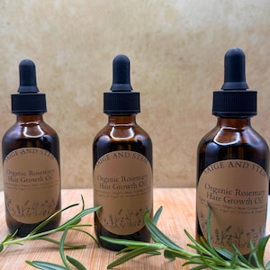 May include: Three amber glass bottles of "Organic Rosemary Hair Growth Oil" with black droppers and brown labels. Fresh rosemary sprigs are scattered around the bottles. The labels read "SAIGE AND STEM".