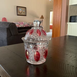 May include: A clear glass candy dish with a lid. The dish has a cut glass design with red accents. The dish is sitting on a dark wood table.