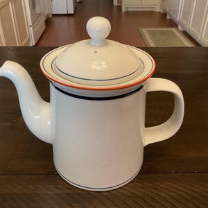 May include: White ceramic teapot with a lid and handle. The lid has a small knob and is trimmed with orange and blue bands. The teapot has a spout and a handle. A classic design for serving tea or coffee.