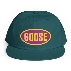 Puede incluir: Una gorra de béisbol de color verde azulado oscuro con un gráfico ovalado central. El óvalo tiene un fondo morado con un borde amarillo y la palabra GOOSE impresa en letras mayúsculas amarillas en el centro. La gorra tiene una corona estructurada.