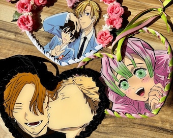 May include: Three heart-shaped wall hangings featuring anime-style illustrations. One displays two characters with blonde and brown hair, adorned with pink roses and pink ribbons. Another shows two characters kissing, framed in black. The third depicts a character with pink and green hair.
