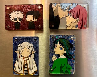 Può includere: Quattro magneti rettangolari in acrilico con illustrazioni di personaggi in stile anime. Uno mostra due figure chibi su uno sfondo rosso glitterato, un altro una coppia faccia a faccia, uno un elfo dai capelli bianchi e l'ultimo un personaggio dai capelli scuri.