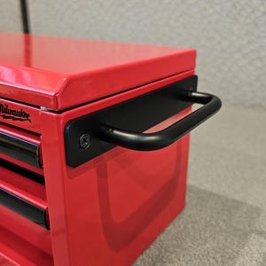 May include: A red metal tool chest with black drawer fronts and a black handle. The Milwaukee logo is visible on the top left corner. The tool chest has three drawers.