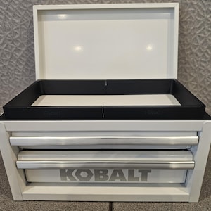 May include: A white metal tool chest with an open lid and two drawers. The drawers have silver handles, and the word "KOBALT" is printed in silver on the bottom drawer. A black tray sits inside the chest.