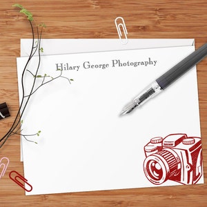 May include: White stationery with the text "Hilary George Photography" printed in black at the top. A red illustration of a vintage camera is at the bottom of the page. A gray fountain pen is lying on the page.