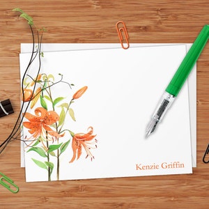 May include: A white card with a watercolor illustration of orange lilies and green leaves. The card has the name "Kenzie Griffin" printed in orange at the bottom.