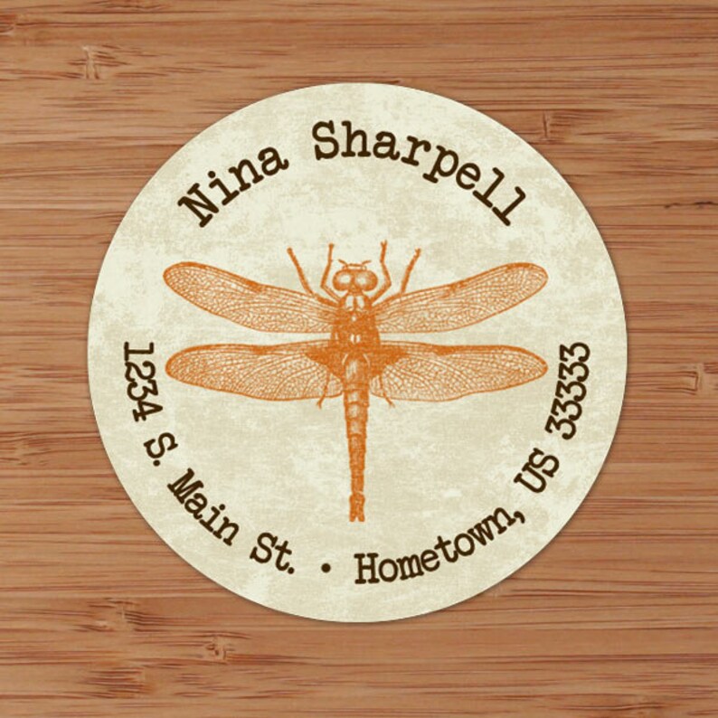 May include: A round, cream-colored sticker with a brown dragonfly illustration in the center. The text "Nina Sharpell" is at the top of the sticker, and the address "1234 S. Main St. Hometown, US 33333" is around the bottom edge.