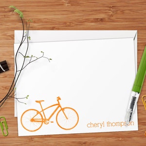 May include: A white card with an orange bicycle illustration and the text "cheryl thompson" at the bottom. The card is on a wooden surface with a green pen and paper clips.