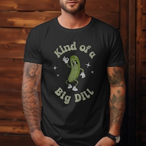 Peut inclure: T-shirt noir avec un dessin animé de cornichon. Le cornichon, portant des gants et des chaussures blancs, fait le signe « ok ». Le texte « Kind of a Big Dill » est arqué au-dessus et en dessous du cornichon. De petits accents d'étoiles complètent le design.