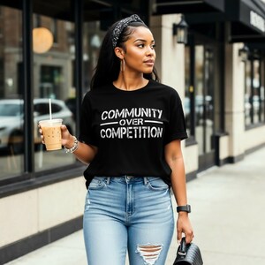 Könnte beinhalten: Schwarzes T-Shirt mit dem weißen Text "COMMUNITY OVER COMPETITION". Die Person trägt eine hellblaue Jeans mit Distressed-Details und ein schwarz-weiß gemustertes Stirnband. Sie hält ein Getränk in einem transparenten Becher.