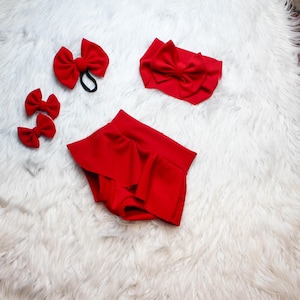 May include: A set of red baby accessories, including a headband with a bow, two small hair bows, a larger hair bow on a black elastic, and a pair of ruffled shorts. The items are arranged on a white, fluffy surface.
