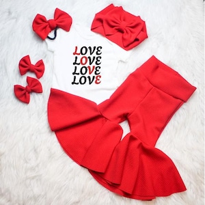 May include: A white onesie with the word "LOVE" in black and red, paired with red flared pants, a headband, and three red bows. The outfit is displayed on a white, fluffy surface.