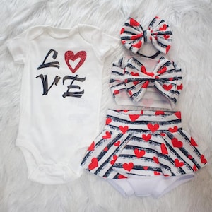 May include: A baby outfit set in white, featuring a bodysuit, skirt, headband, and bow. The bodysuit has the word "LOVE" in black with a red glitter heart. The skirt, headband, and bow have a black and white striped pattern with red hearts.