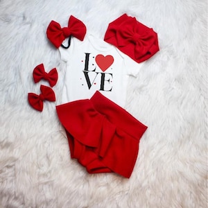 May include: A baby outfit set featuring a white onesie with the word "LOVE" and a red heart, paired with red shorts, bows, and a headband. The set is arranged on a white, fluffy surface.