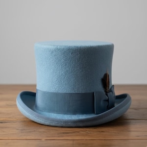 May include: A light blue top hat with a matching ribbon and a small feather accent. The hat has a rounded crown and a brim. The hat is sitting on a wooden surface. This formal hat is suitable for special occasions.