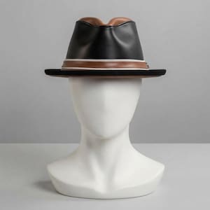 Könnte beinhalten: Ein Fedora-Hut mit schwarzer Krone und breiter schwarzer Krempe. Der Hut ist mit einem hellbraunen Lederband akzentuiert, das von dünnen weißen Streifen gesäumt wird. Das Design umfasst eine eingedrückte Krone und eine strukturierte, flache Krempe.
