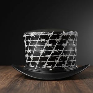 May include: A black top hat with a white barbed wire pattern. The hat has a wide brim and is decorated with silver studs. The hat is made of leather and sits on a wooden surface.