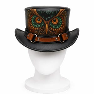 May include: A black top hat featuring a detailed hand-painted owl face on the front crown. The owl has large orange eyes and green feathers. A brown leather strap with silver-toned metal rings and rivets encircles the base of the crown.