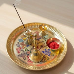 May include: A gold-colored metal tray with a floral design, holding a small bowl of red powder, a lit candle, a bell, and an incense burner with a lit incense stick. The tray is round with a decorative edge.