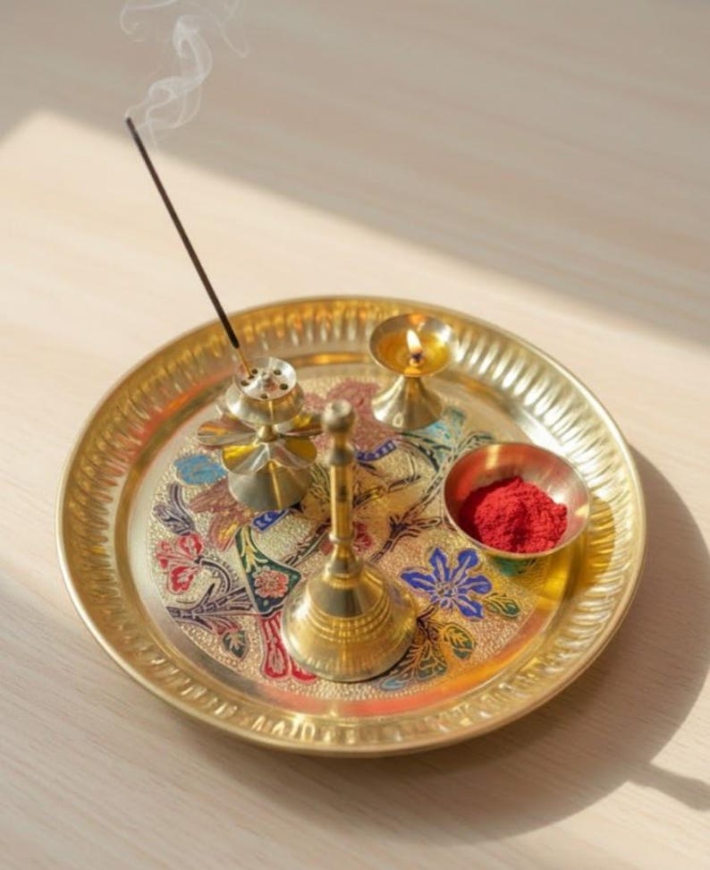 May include: A gold-colored metal tray with a floral design, holding a small bowl of red powder, a lit candle, a bell, and an incense burner with a lit incense stick. The tray is round with a decorative edge.