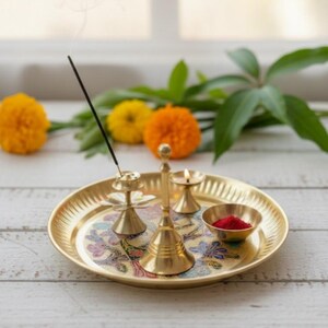 May include: A gold-colored brass puja thali set with a floral design. The set includes a bell, incense holder, small bowl with red powder, and a candle holder with a lit candle. Incense smoke rises from the incense holder. Marigold flowers are in the background.