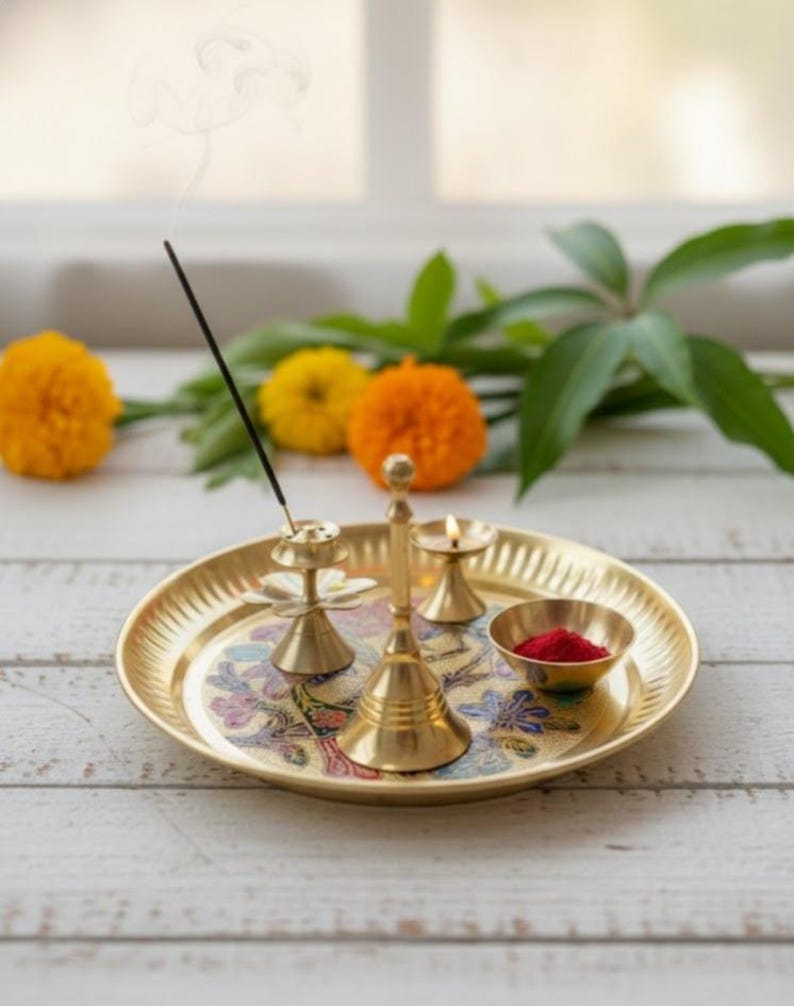 May include: A gold-colored brass puja thali set with a floral design. The set includes a bell, incense holder, small bowl with red powder, and a candle holder with a lit candle. Incense smoke rises from the incense holder. Marigold flowers are in the background.