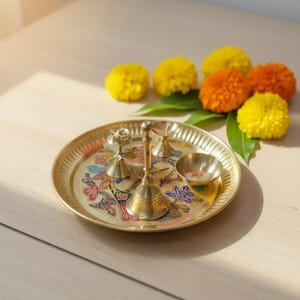 May include: A brass puja thali with floral designs and small brass bells and bowls. The thali is surrounded by yellow and orange marigold flowers and green leaves, creating a vibrant and spiritual display.
