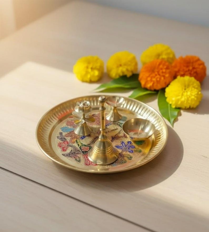 May include: A brass puja thali with floral designs and small brass bells and bowls. The thali is surrounded by yellow and orange marigold flowers and green leaves, creating a vibrant and spiritual display.