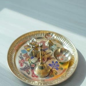 May include: A gold-colored metal puja thali set with a floral design. The set includes a small bell, a bowl, a small incense holder, and two small candle holders. The thali is round with a decorative edge.