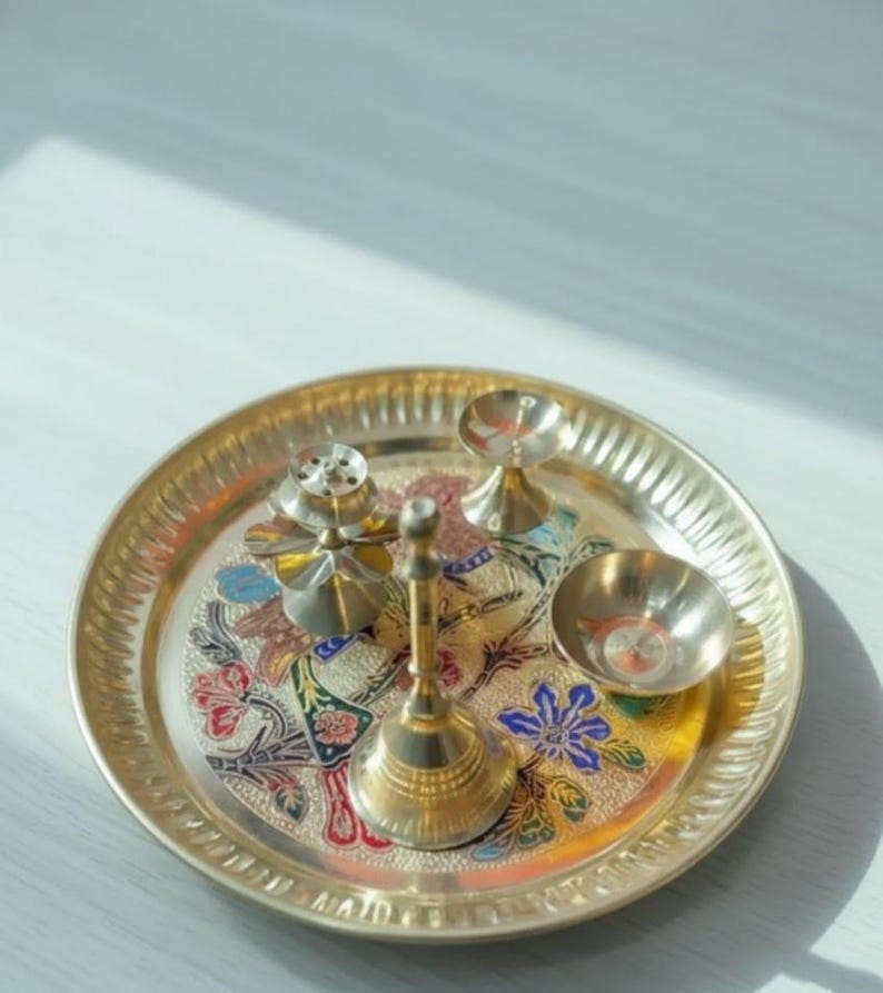 May include: A gold-colored metal puja thali set with a floral design. The set includes a small bell, a bowl, a small incense holder, and two small candle holders. The thali is round with a decorative edge.