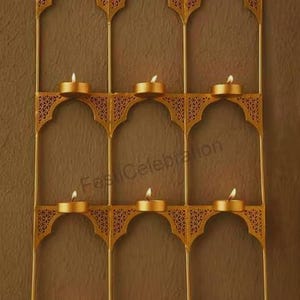 May include: A gold-toned metal wall-mounted candle holder featuring a grid of nine arched compartments. Each arch contains a small, lit tea light candle. The frame has intricate, laser-cut filigree detailing in the upper corners of each arch.