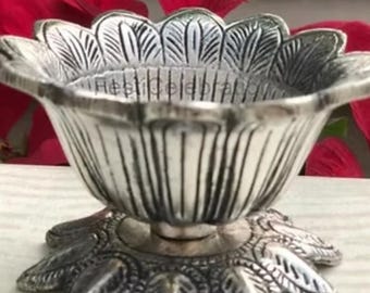 May include: A silver-colored, decorative bowl with a floral design. The bowl has a petal-shaped rim and a base with leaf-shaped details. The background features red flowers.