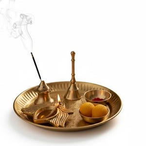 May include: A golden brass puja thali with various items for worship. Includes a lit oil lamp, incense, a small bell, two bowls with red powder and yellow sweets, and a smoking incense stick. The items are arranged on a round, textured tray.