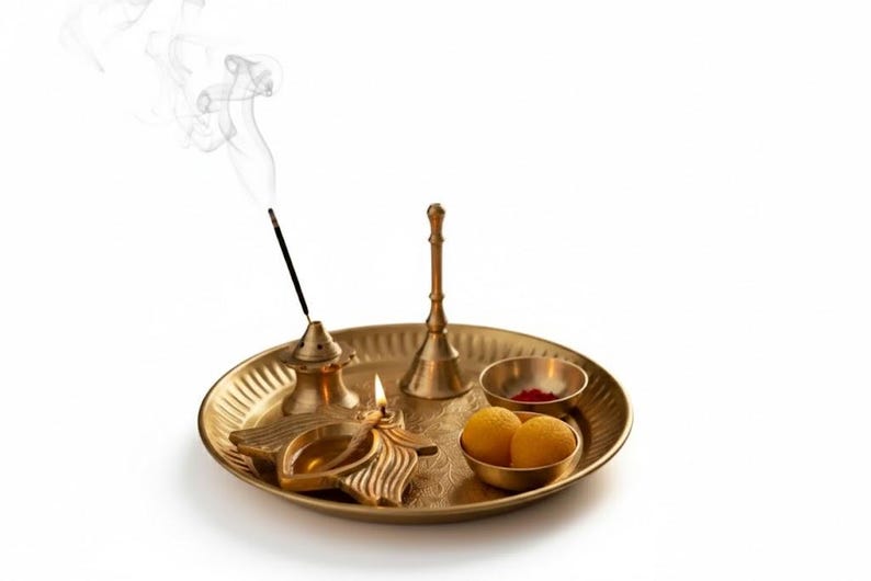 May include: A golden brass puja thali with various items for worship. Includes a lit oil lamp, incense, a small bell, two bowls with red powder and yellow sweets, and a smoking incense stick. The items are arranged on a round, textured tray.